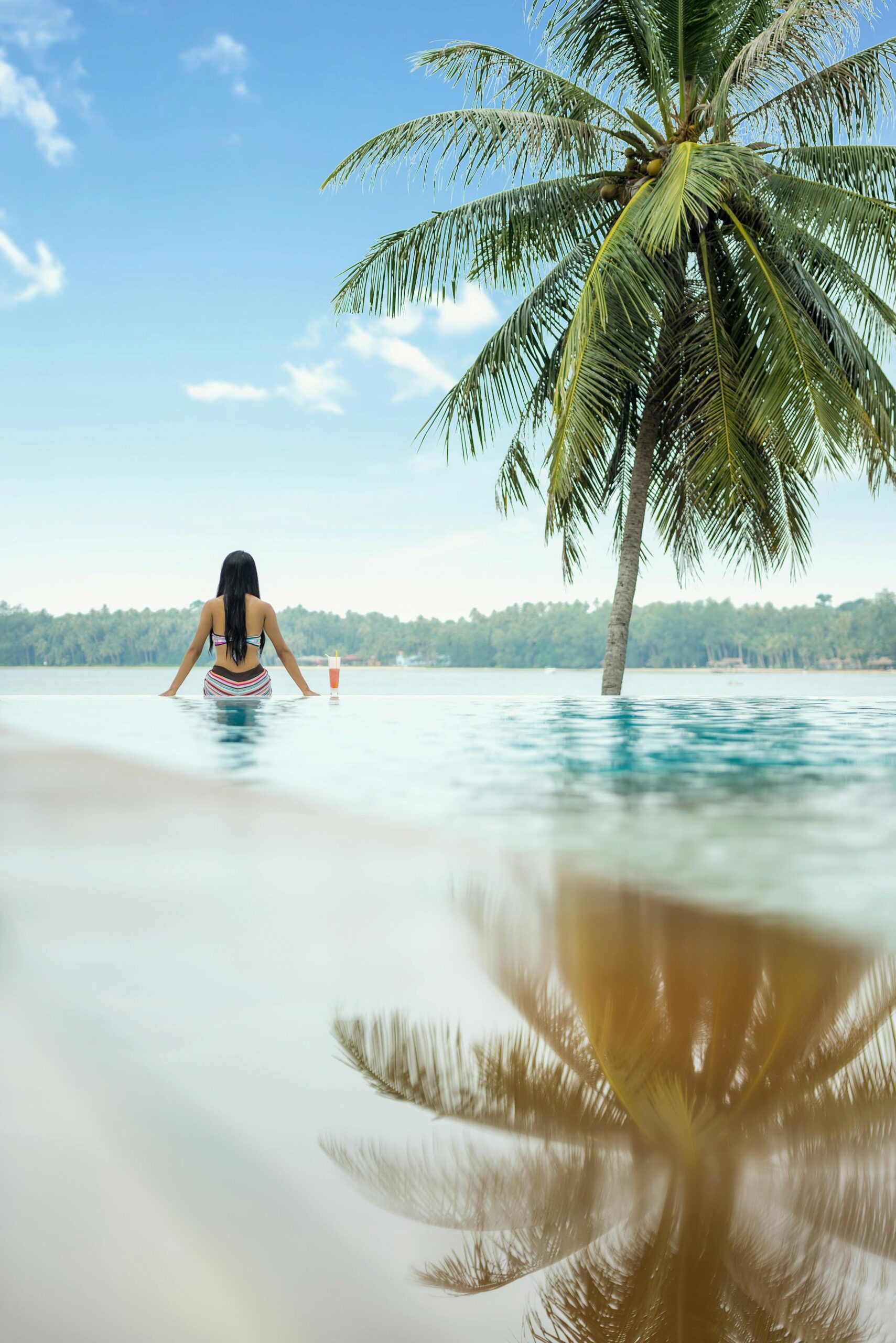 Woman relaxing in an infinity pool with a palm tree and ocean view.